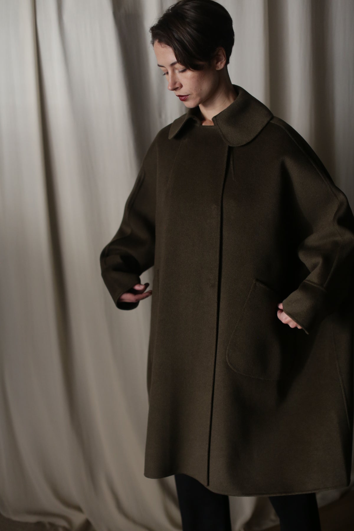 Wearing the Kyoto | Olive coat against a neutral draped background, a person with short dark hair looks down as they tuck their hands into sizable pockets. Crafted from double-faced cashmere, the coat's dramatic silhouette stands out.