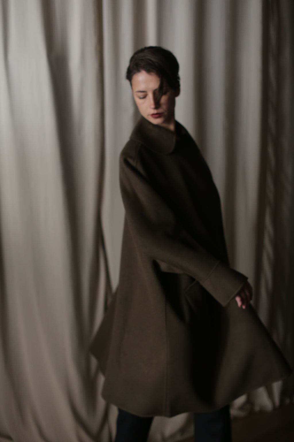 In dim light, a person with dark hair and a Kyoto | Olive coat stands lightly turned before a cream curtain, casting soft shadows. The lighting accentuates the voluminous coat's shape, creating a dramatic silhouette and moody ambiance.