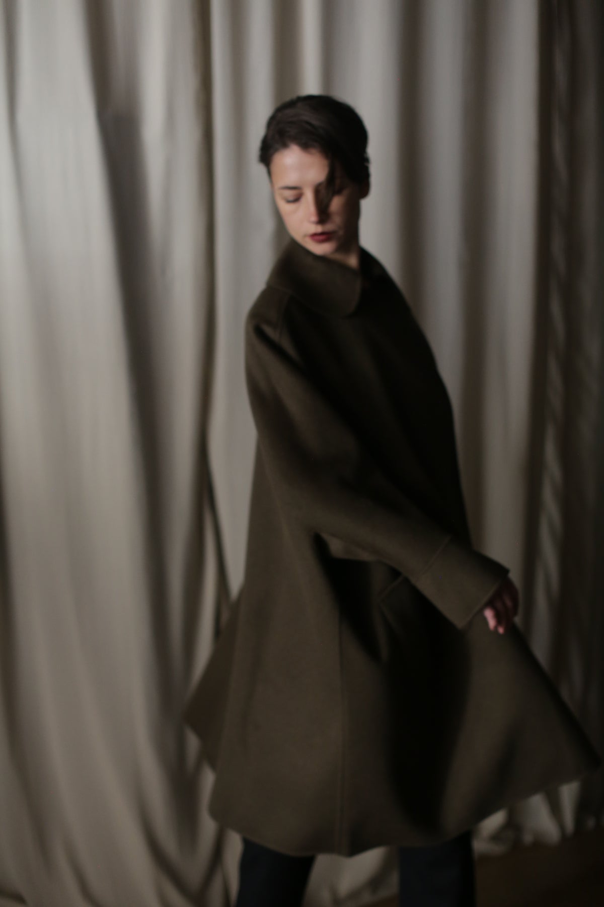 In dim light, a person with dark hair and a Kyoto | Olive coat stands lightly turned before a cream curtain, casting soft shadows. The lighting accentuates the voluminous coat's shape, creating a dramatic silhouette and moody ambiance.