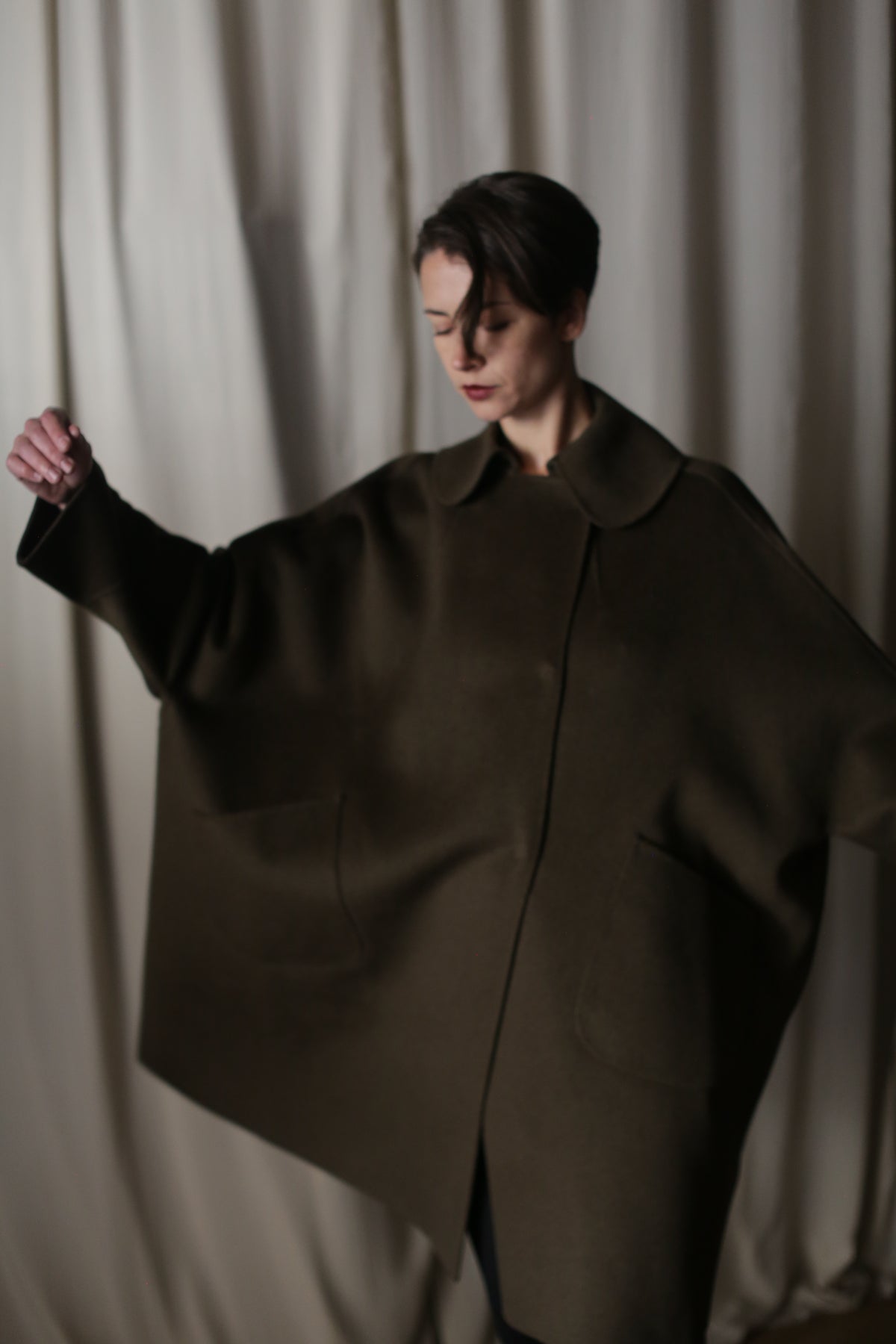 A person stands with a neutral expression against a draped curtain backdrop, the oversized Kyoto Olive coat creating a dramatic silhouette as they hold their arms outstretched.