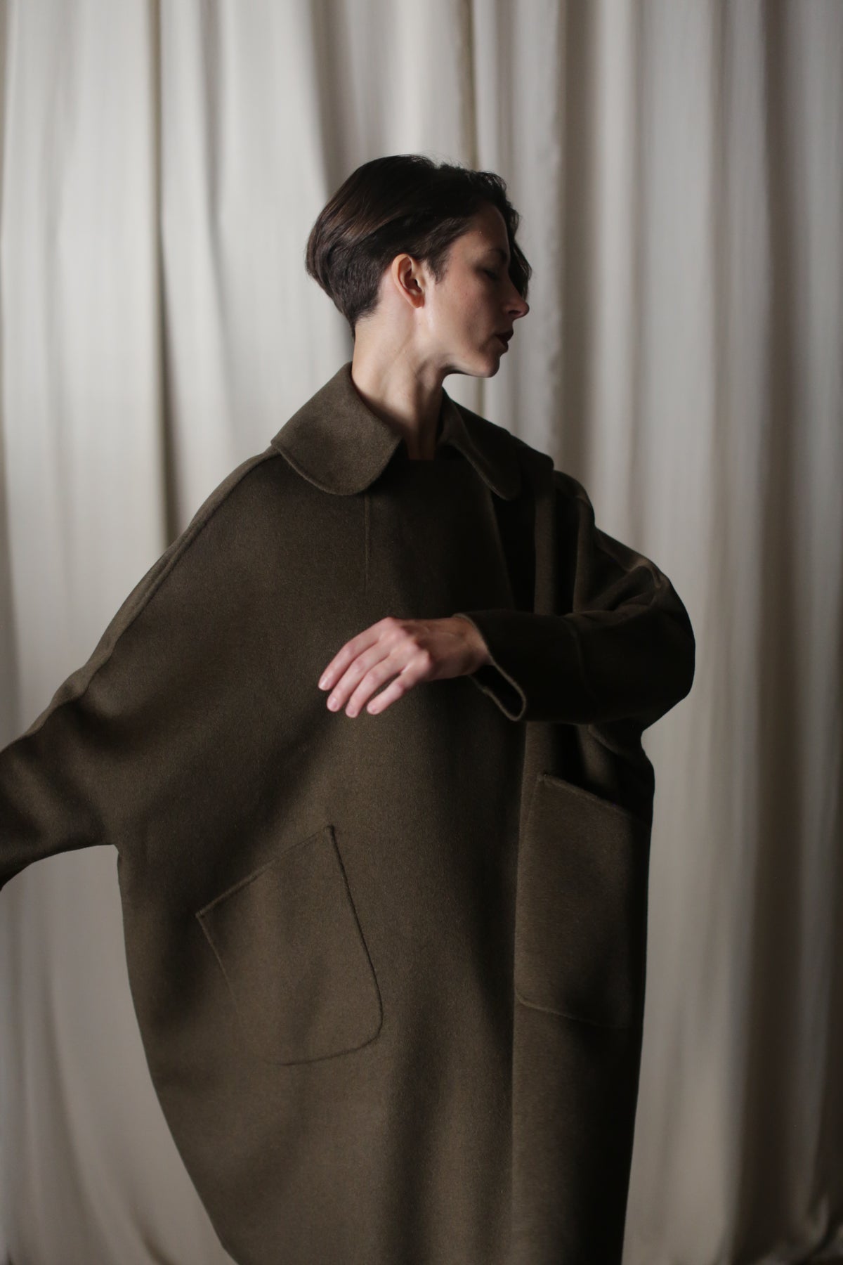 A person with short hair wears the Kyoto | Olive oversized coat in dark olive double-faced cashmere, featuring large pockets. They stand before a cream-colored curtain, creating a dramatic silhouette with one arm raised and head turned to the side.