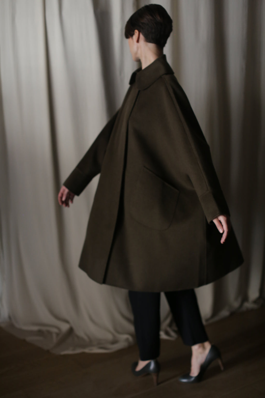 A person with short hair models the Kyoto | Olive oversized coat, featuring a dramatic silhouette and large pockets, as they walk leftward. They pair it with black pants and metallic heels, set against a backdrop of beige curtains that create a soft, elegant atmosphere.