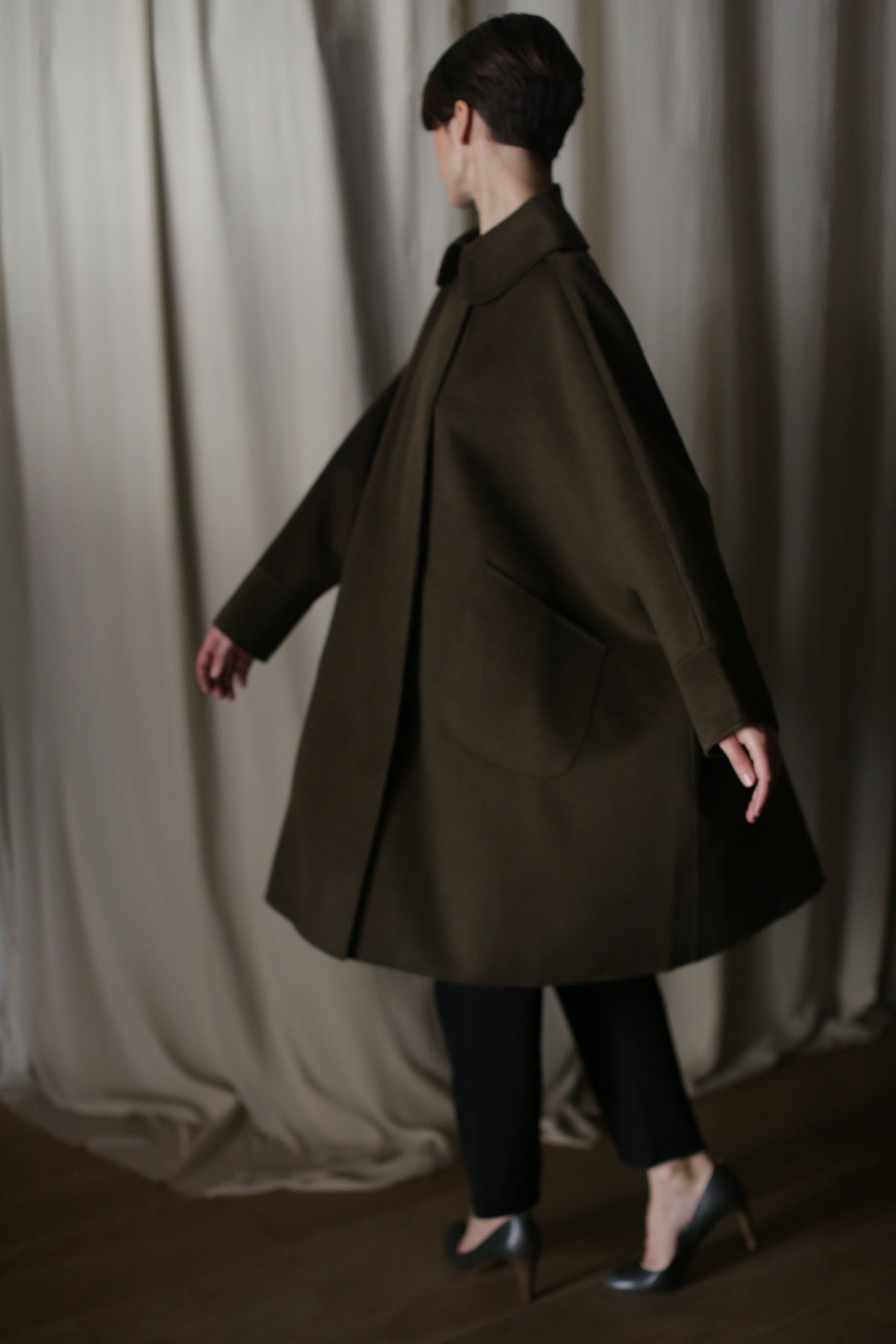 A person with short hair models the Kyoto | Olive oversized coat, featuring a dramatic silhouette and large pockets, as they walk leftward. They pair it with black pants and metallic heels, set against a backdrop of beige curtains that create a soft, elegant atmosphere.