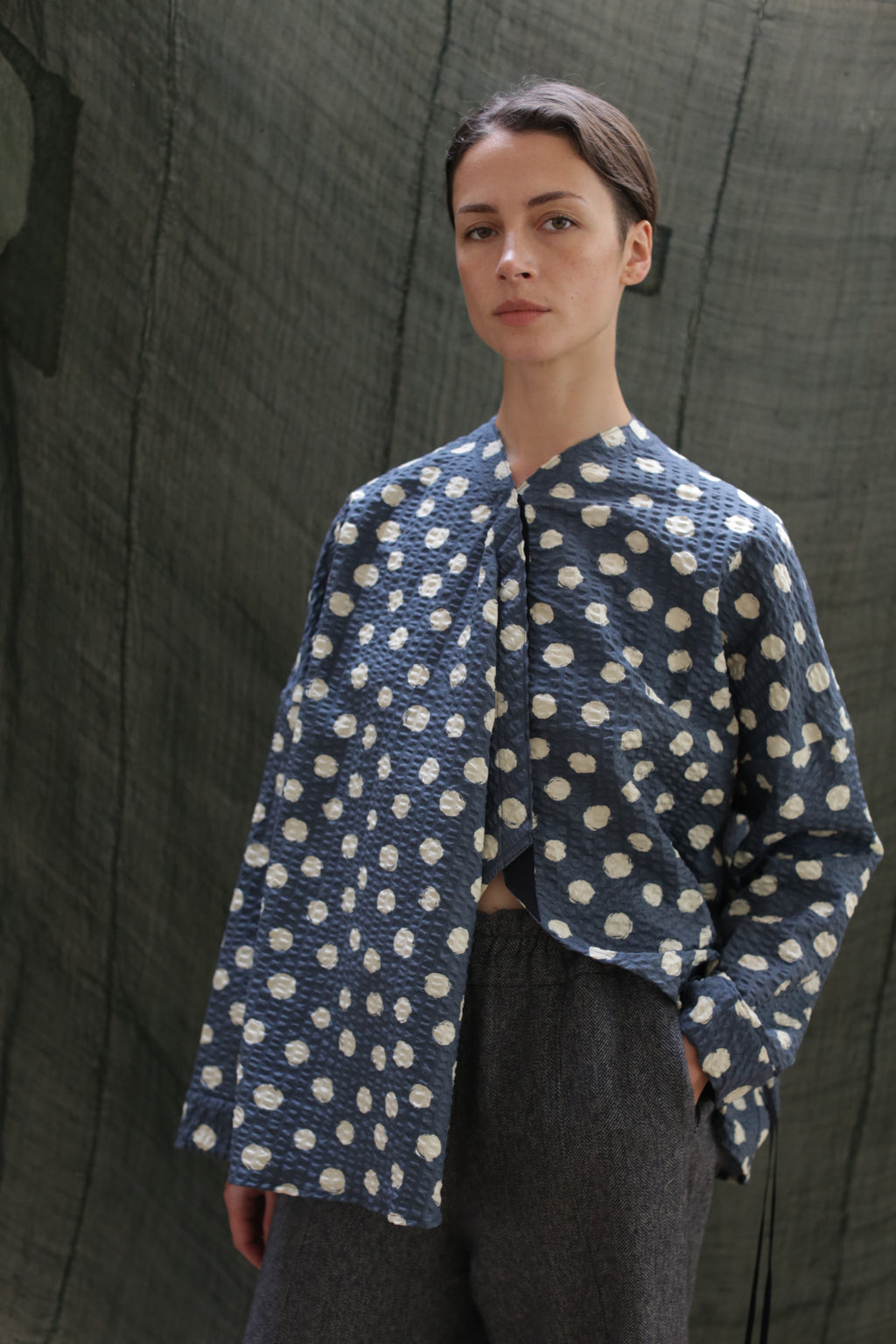 A person with short hair wears the Qipao Blouse | Polkadot Seersucker Indigo and loose gray pants, standing before a sheer dark green backdrop and looking at the camera with a neutral expression.
