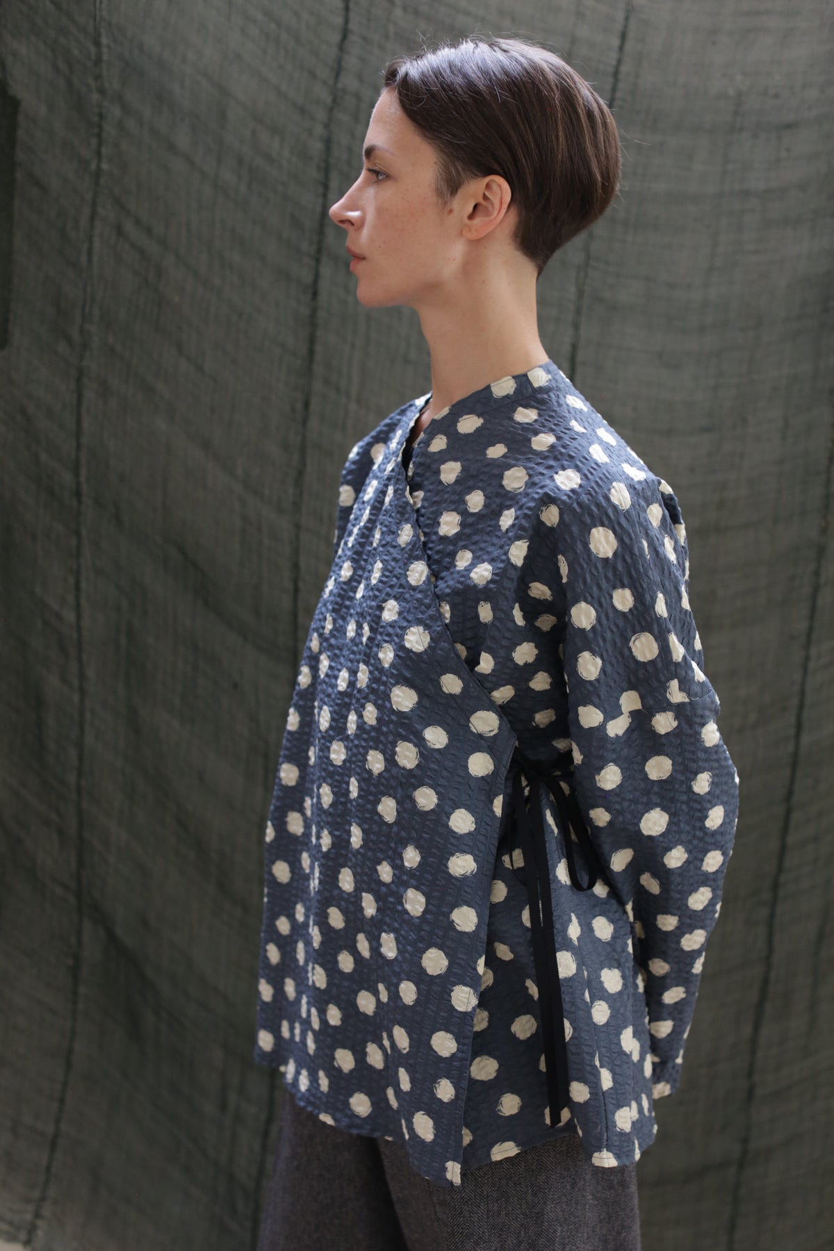 A person with short brown hair stands in profile against a dark fabric backdrop, wearing the Qipao Blouse | Polkadot Seersucker Indigo, a loose blue top with white polka dots and their hands behind their back.