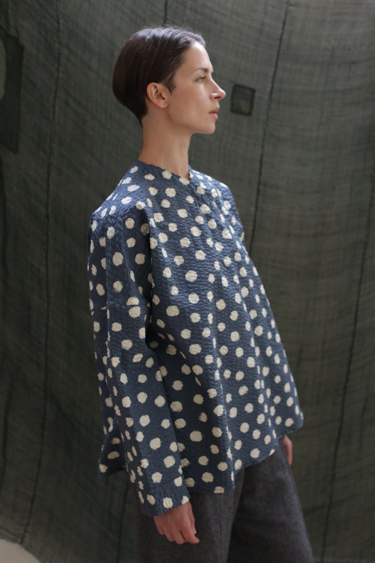 A person with short brown hair wears the Qipao Blouse | Polkadot Seersucker Indigo—an indigo, loose-fitting long-sleeve top with large white polkadots—paired with gray pants, standing in profile against a textured greenish-gray backdrop.