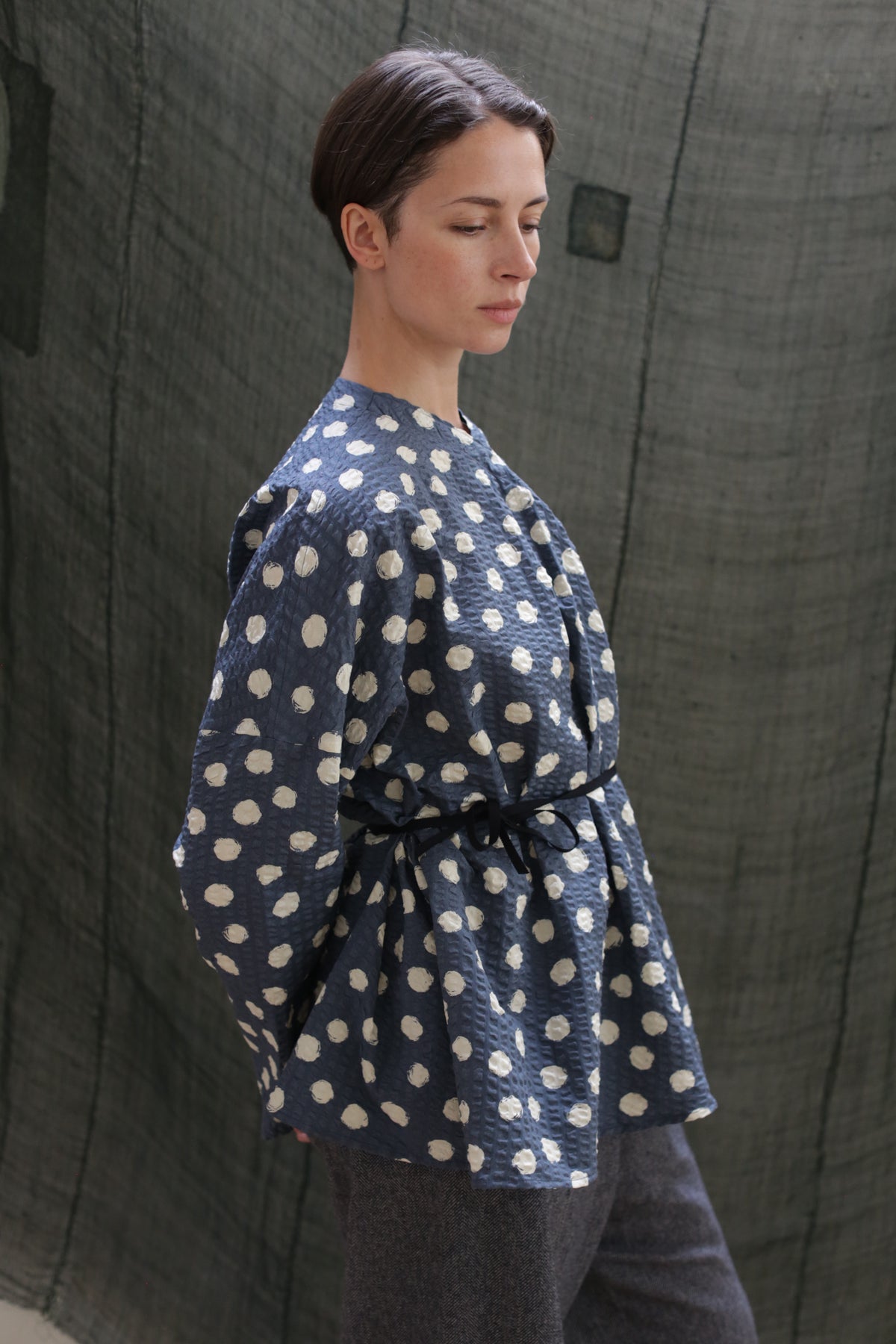 A person with short dark hair stands sideways, looking down, wearing the Qipao Blouse | Polkadot Seersucker Indigo belted at the waist with gray trousers. The background features textured dark green fabric.