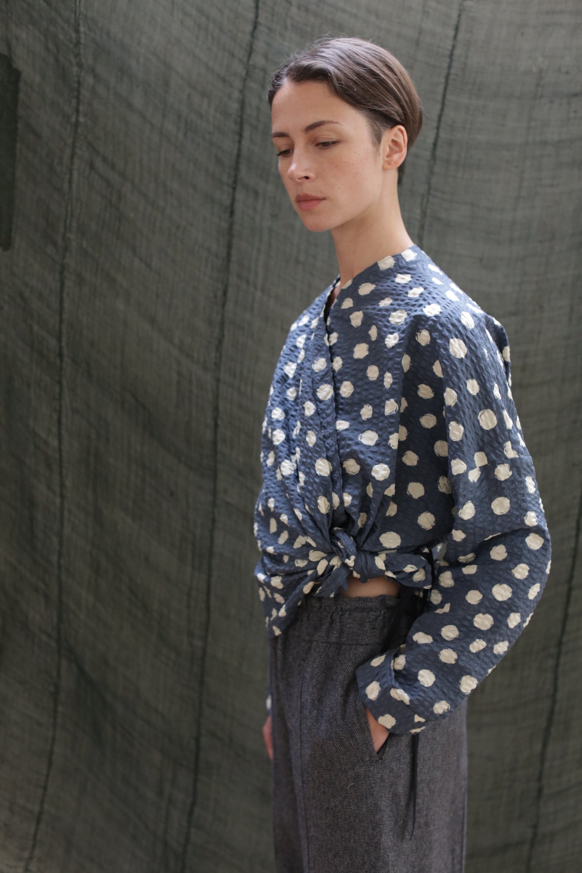 A person with short brown hair wears the Qipao Blouse | Polkadot Seersucker Indigo, tied at the waist, and gray high-waisted pants, standing in profile against a textured green fabric backdrop.