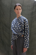 A woman in the Qipao Blouse | Polkadot Seersucker Indigo, featuring a side knot, and gray pants stands before a textured dark green backdrop, looking to the side with a neutral expression.