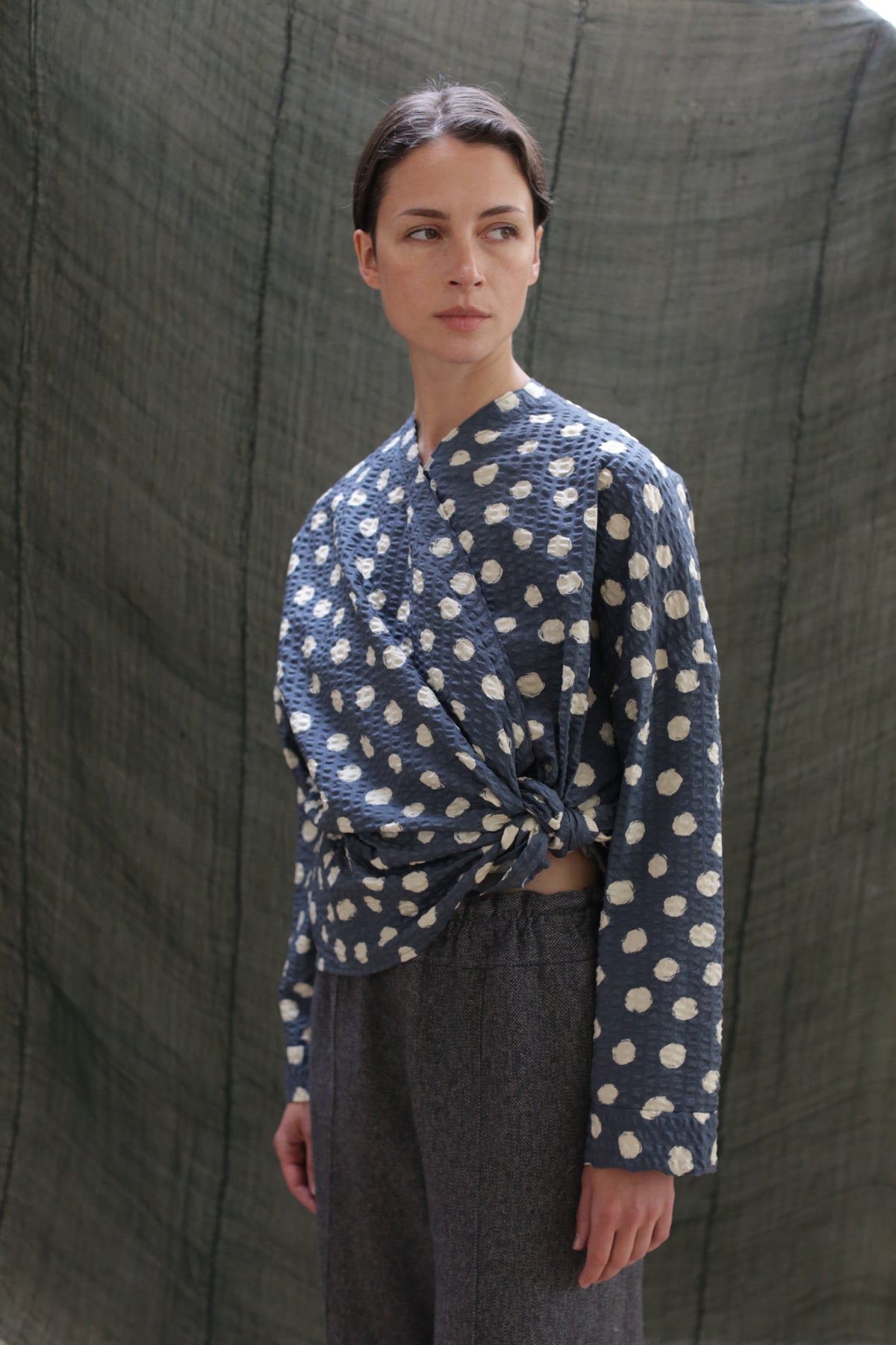 A woman in the Qipao Blouse | Polkadot Seersucker Indigo, featuring a side knot, and gray pants stands before a textured dark green backdrop, looking to the side with a neutral expression.