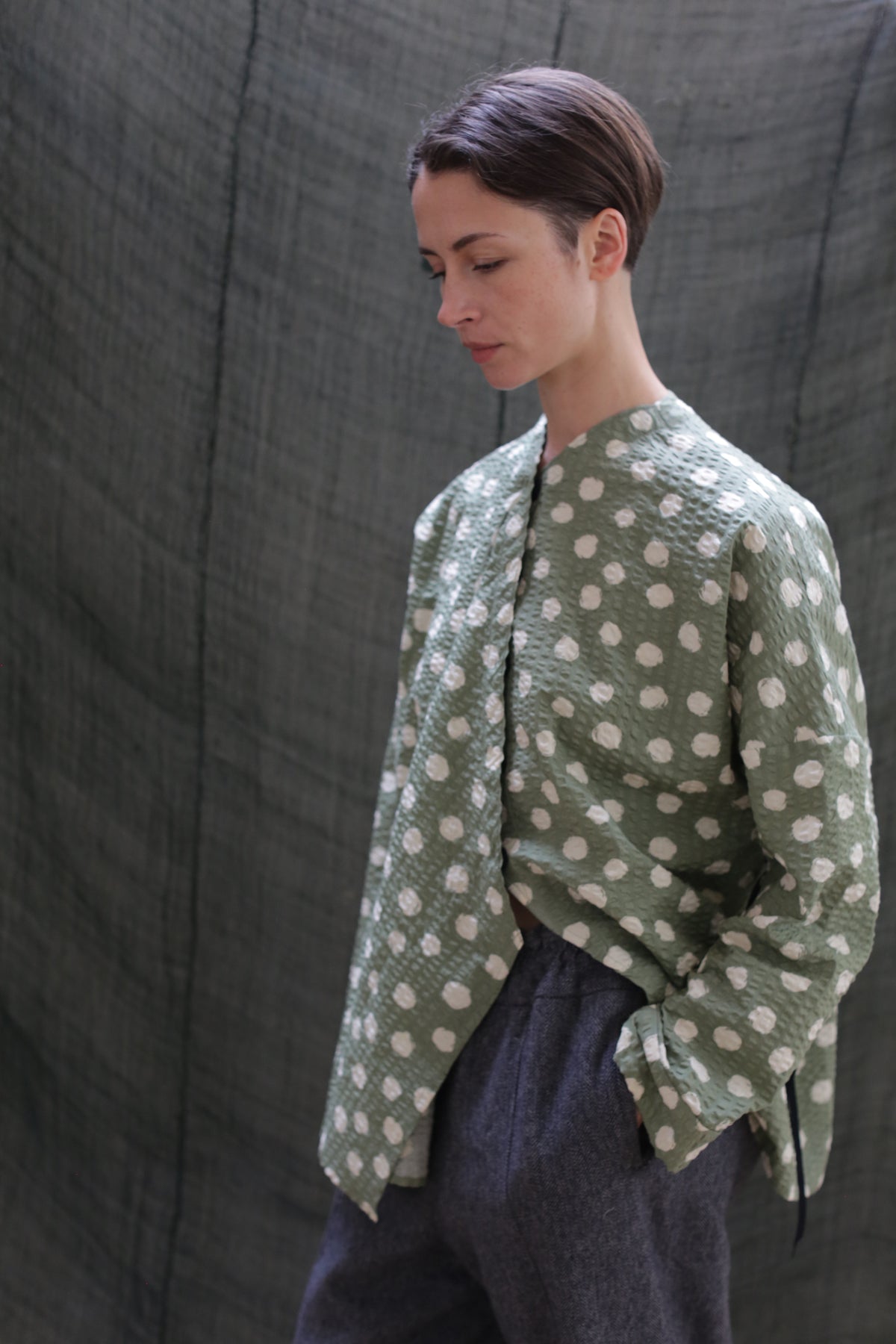 A person with short brown hair wears the Qipao Blouse | Polkadot Seersucker Moss and gray trousers, standing before a textured dark green fabric backdrop, gazing down with one hand in their pocket.