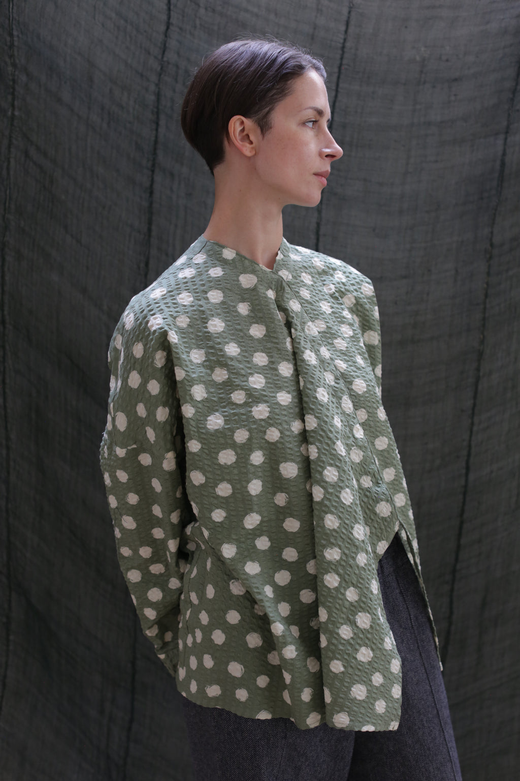 A person with short dark hair, wearing the Qipao Blouse in Polkadot Seersucker Moss and dark pants, stands before a textured dark fabric backdrop, gazing to the side.