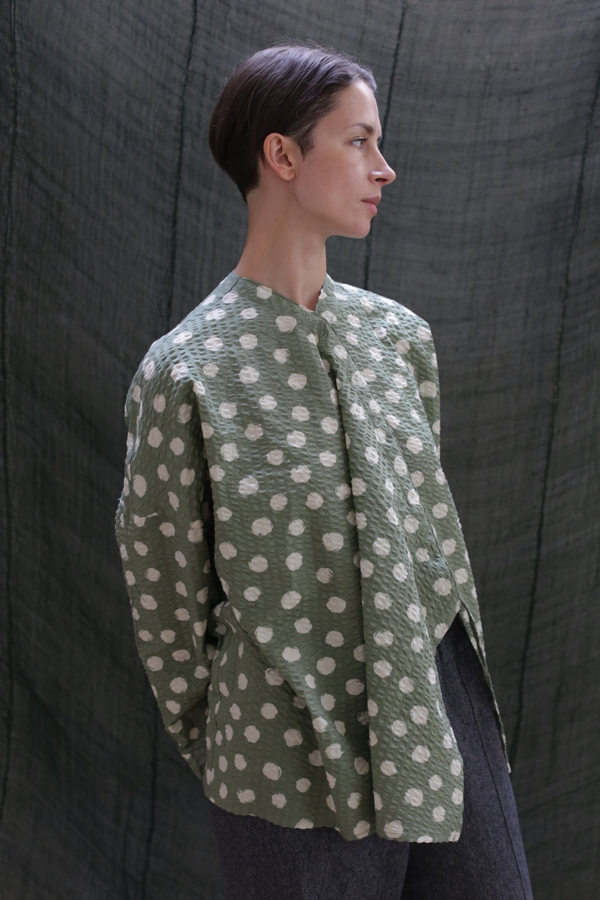 A person with short dark hair, wearing the Qipao Blouse in Polkadot Seersucker Moss and dark pants, stands before a textured dark fabric backdrop, gazing to the side.