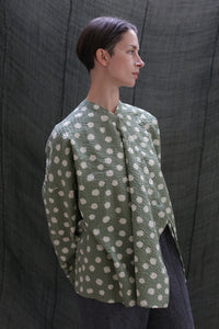 A person with short dark hair, wearing the Qipao Blouse in Polkadot Seersucker Moss and dark pants, stands before a textured dark fabric backdrop, gazing to the side.
