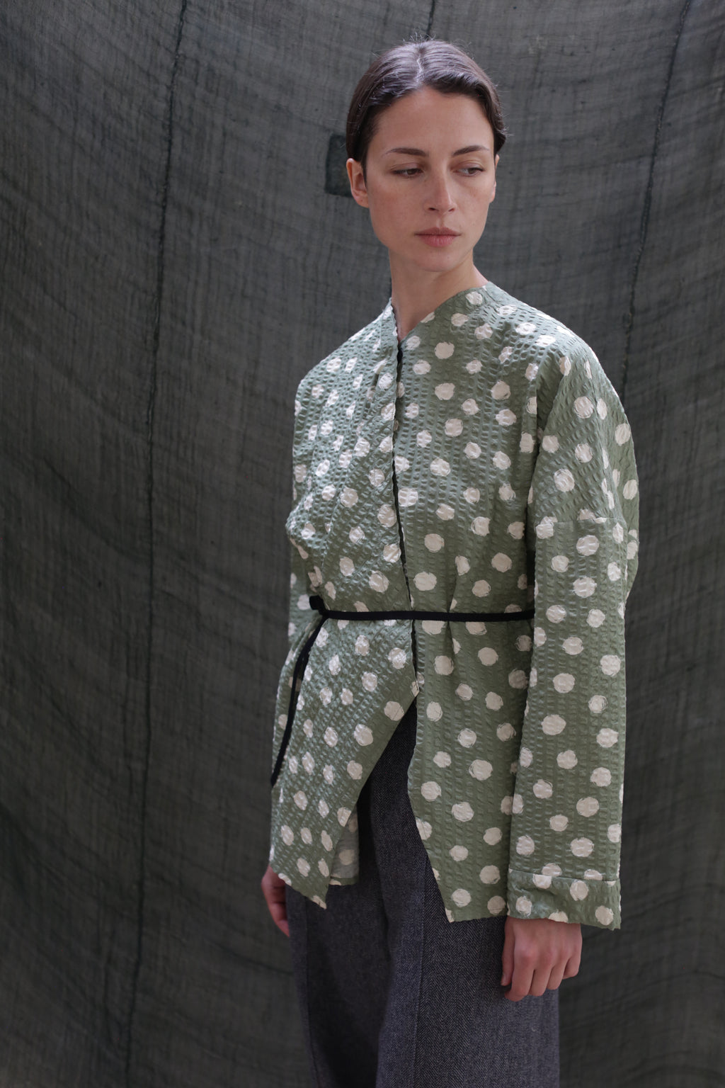 A woman in the Qipao Blouse | Polkadot Seersucker Moss, belted at the waist and paired with gray pants, stands before a textured dark green backdrop, gazing calmly to her left.