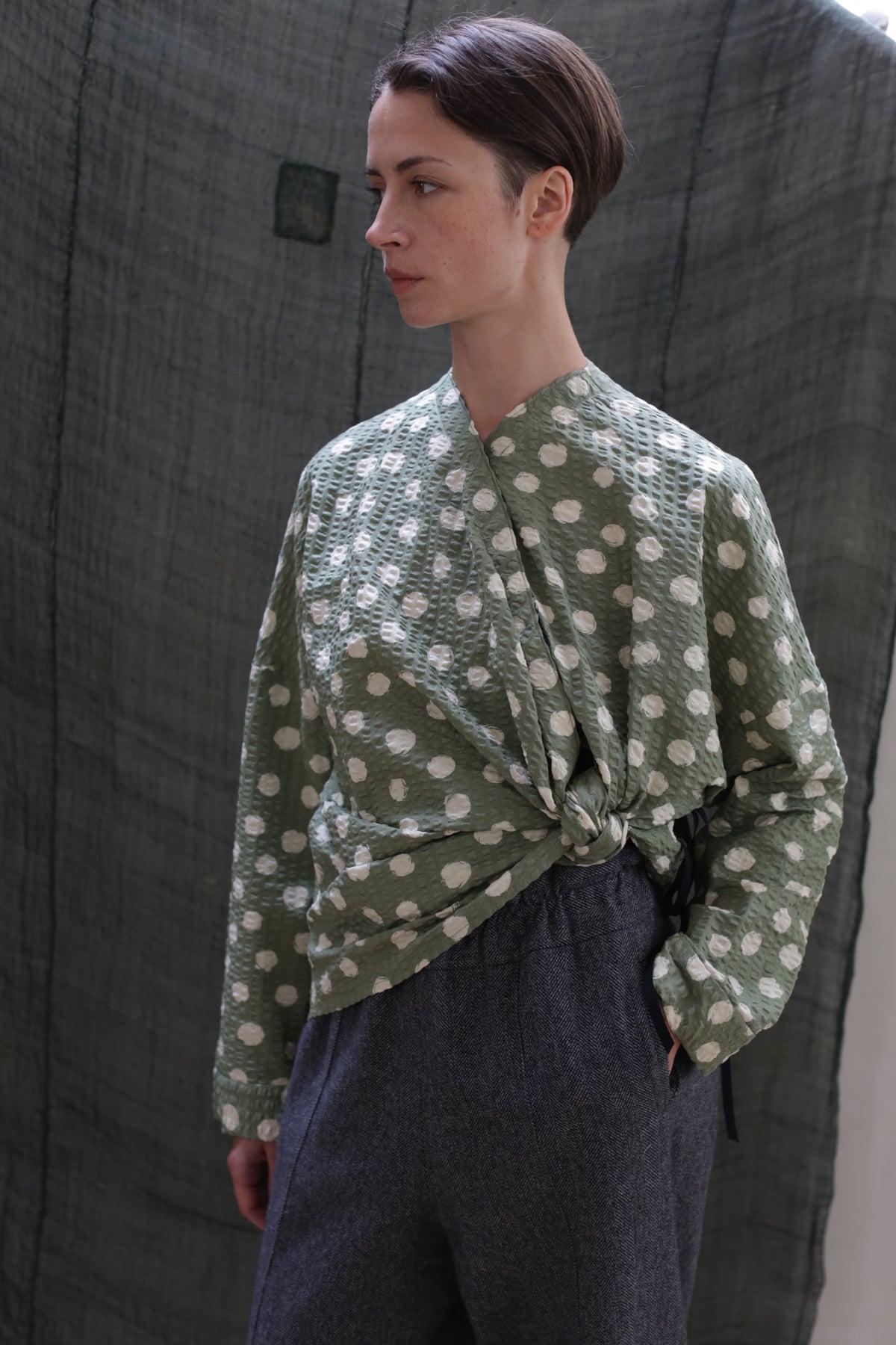A woman with short brown hair wears the Qipao Blouse | Polkadot Seersucker Moss and gray pants, standing before a textured dark green fabric backdrop, gazing to the side.