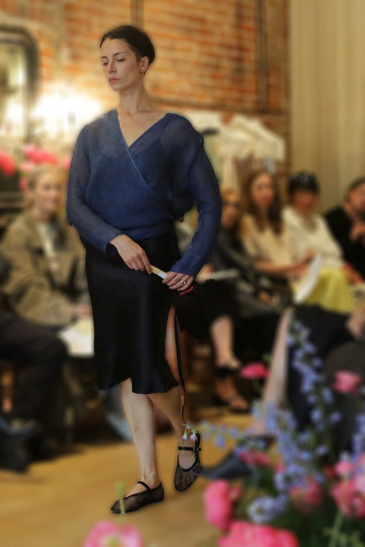 A woman models the X Blouse | Linen Ai Dye and a black skirt, walking with a cane on the runway. Attentive guests watch in a warmly lit room decorated with flowers in the foreground.