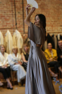 A woman in a flowing Kimono Hand Pleated Dress | Silk Charmeuse Lilac holds a white fan above her head as she walks past a seated audience, with a blurred brick wall and hanging garments in the background.