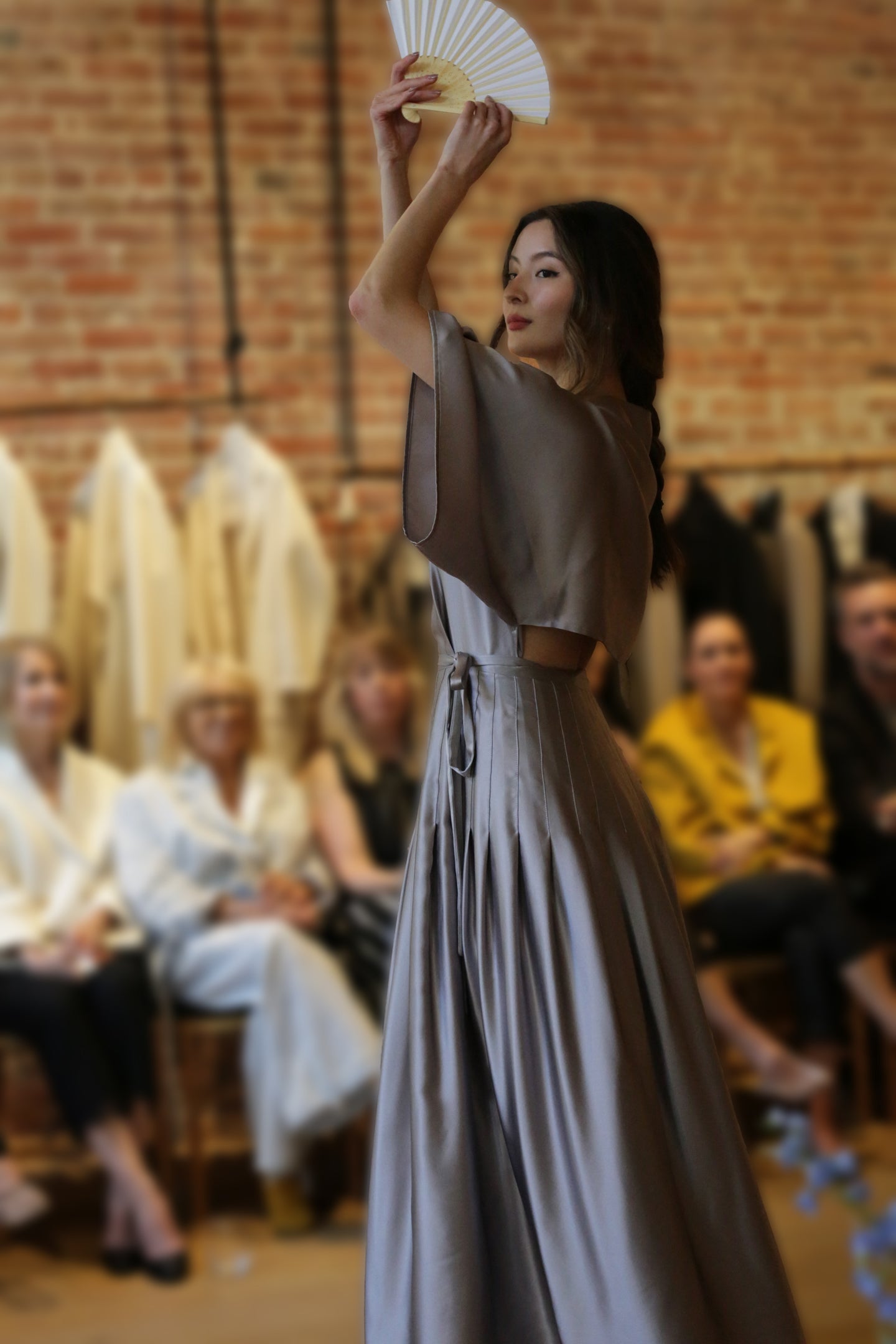 A woman in a flowing Kimono Hand Pleated Dress | Silk Charmeuse Lilac holds a white fan above her head as she walks past a seated audience, with a blurred brick wall and hanging garments in the background.