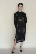 A person with short dark hair wears the sheer black floral Uragiri Dress over a nude slip, paired with black heels, standing against a plain light background with hands behind their back.