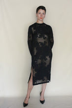 Load image into Gallery viewer, A person with short dark hair wears a sheer black Crewneck Dress | Silk Uragiri Black, featuring floral patterns and a side slit that reveals a nude slip. They stand against a plain light background, looking confidently at the camera.