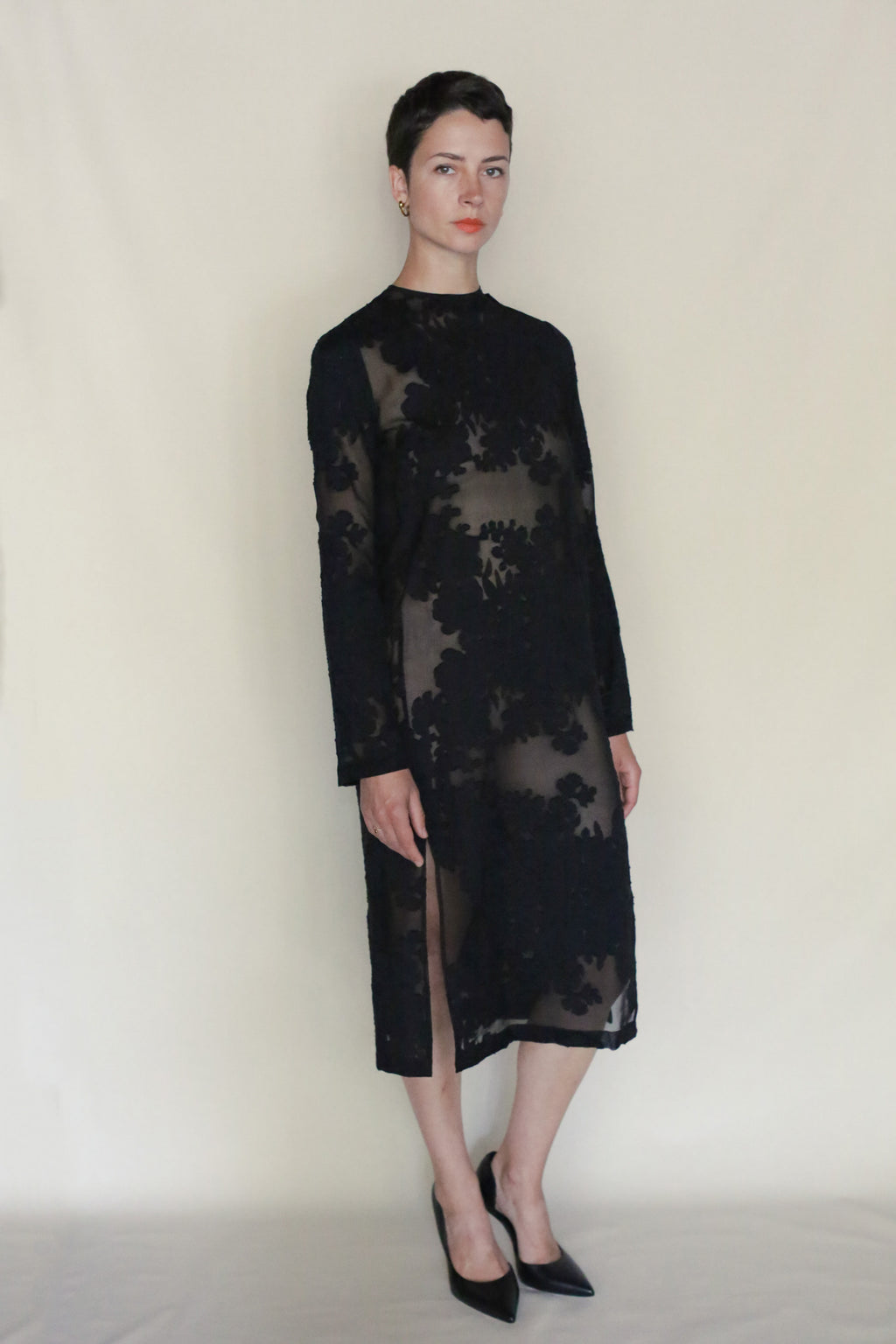 A person stands against a plain background wearing the Uragiri Dress, a black long-sleeve sheer crewneck dress with floral patterns and a side slit, paired with black pointed-toe heels.