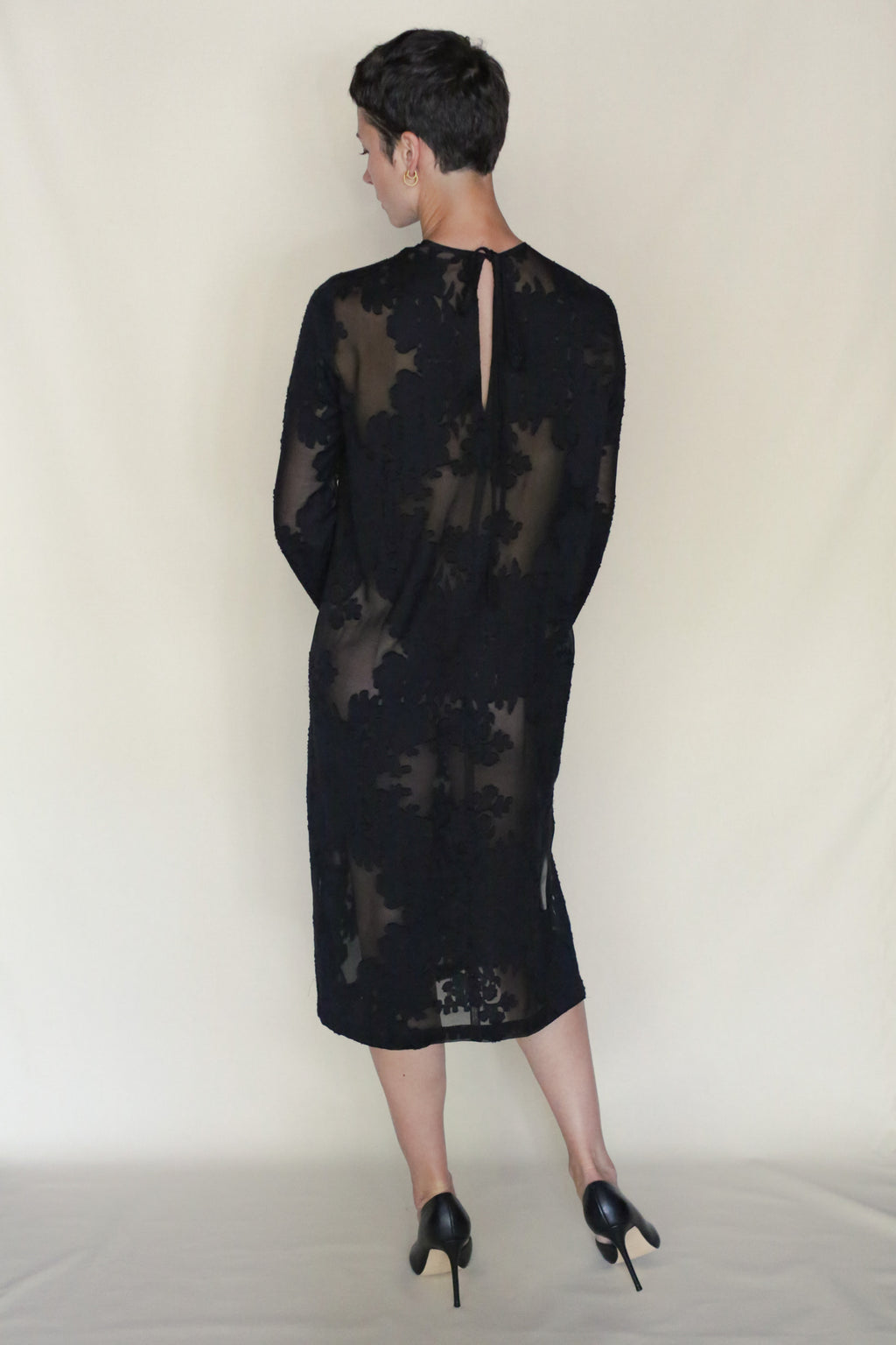 A person with short dark hair stands facing away, wearing a sheer black Uragiri Dress with floral patterns and black high heels, set against a plain light background.