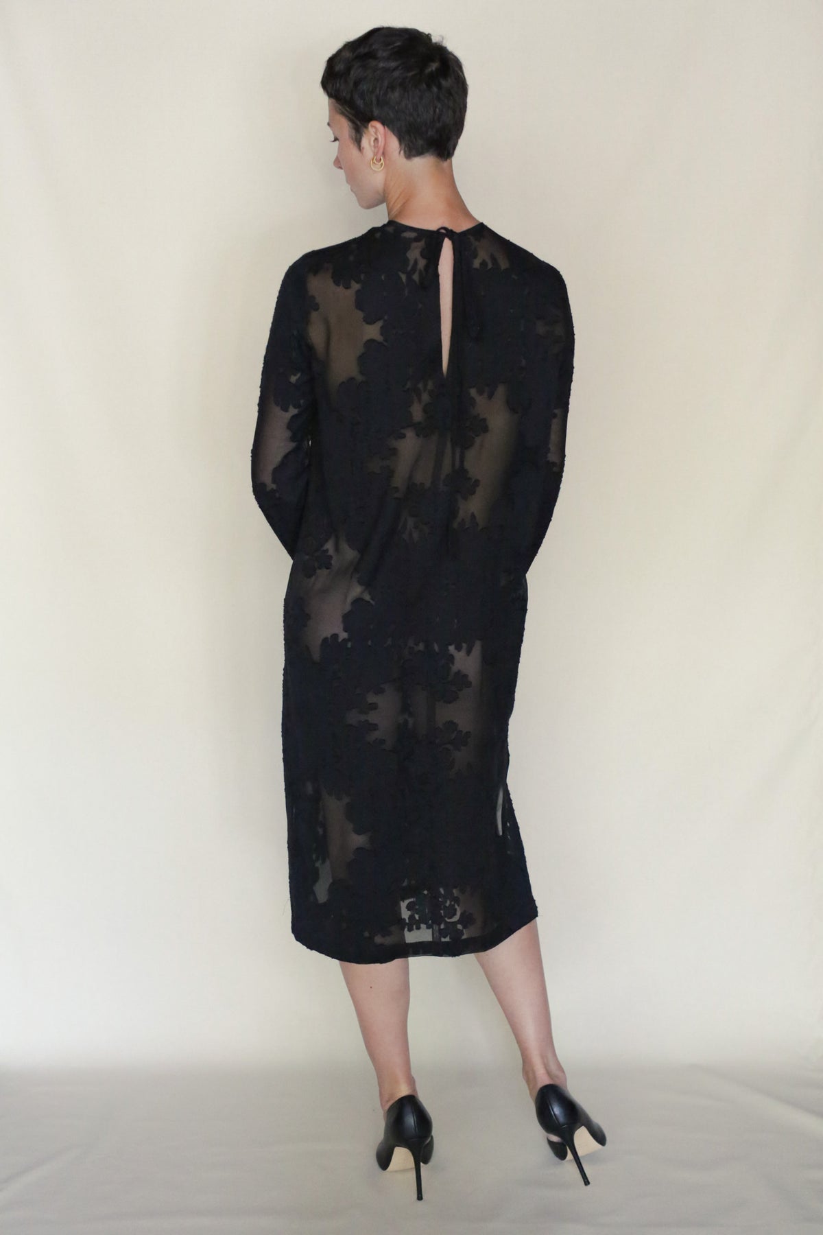A person with short dark hair stands facing away, wearing a sheer black Uragiri Dress with floral patterns and black high heels, set against a plain light background.