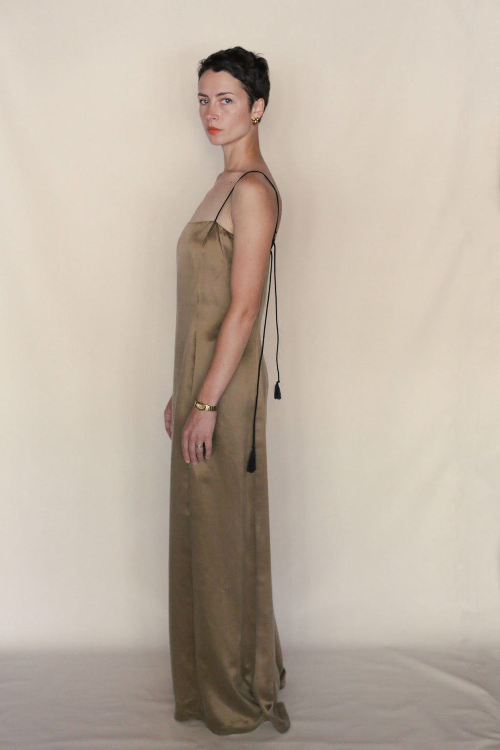 A person with short dark hair wears a sleeveless, floor-length Silk Cord Dress with black spaghetti straps, standing sideways against a plain beige background. They accessorize with gold earrings and a bracelet.