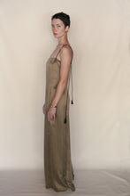 Load image into Gallery viewer, A person with short dark hair stands sideways, wearing the handmade Silk Cord Dress | Gold featuring thin black straps and tassels. The plain beige background highlights the dress’s elegant San Francisco style.