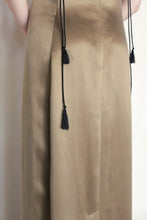 Load image into Gallery viewer, A close-up of a person wearing the Silk Cord Dress | Gold, featuring two black tassel cords at the front. The image shows only the lower torso and arms, emphasizing this handmade San Francisco-style garment.