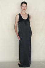 Load image into Gallery viewer, A person with short dark hair stands against a plain light background, wearing the Ink Stone Dress—a long, sleeveless black cupro dress with a scoop neckline and side pockets. This handmade piece from San Francisco is styled simply with hands in the pockets.