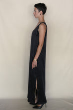 Load image into Gallery viewer, A person with short dark hair stands in profile against a plain background, wearing the handmade Ink Stone Dress—a sleeveless, long black cupro dress with a side slit—paired with black heels and a bracelet on their left wrist.