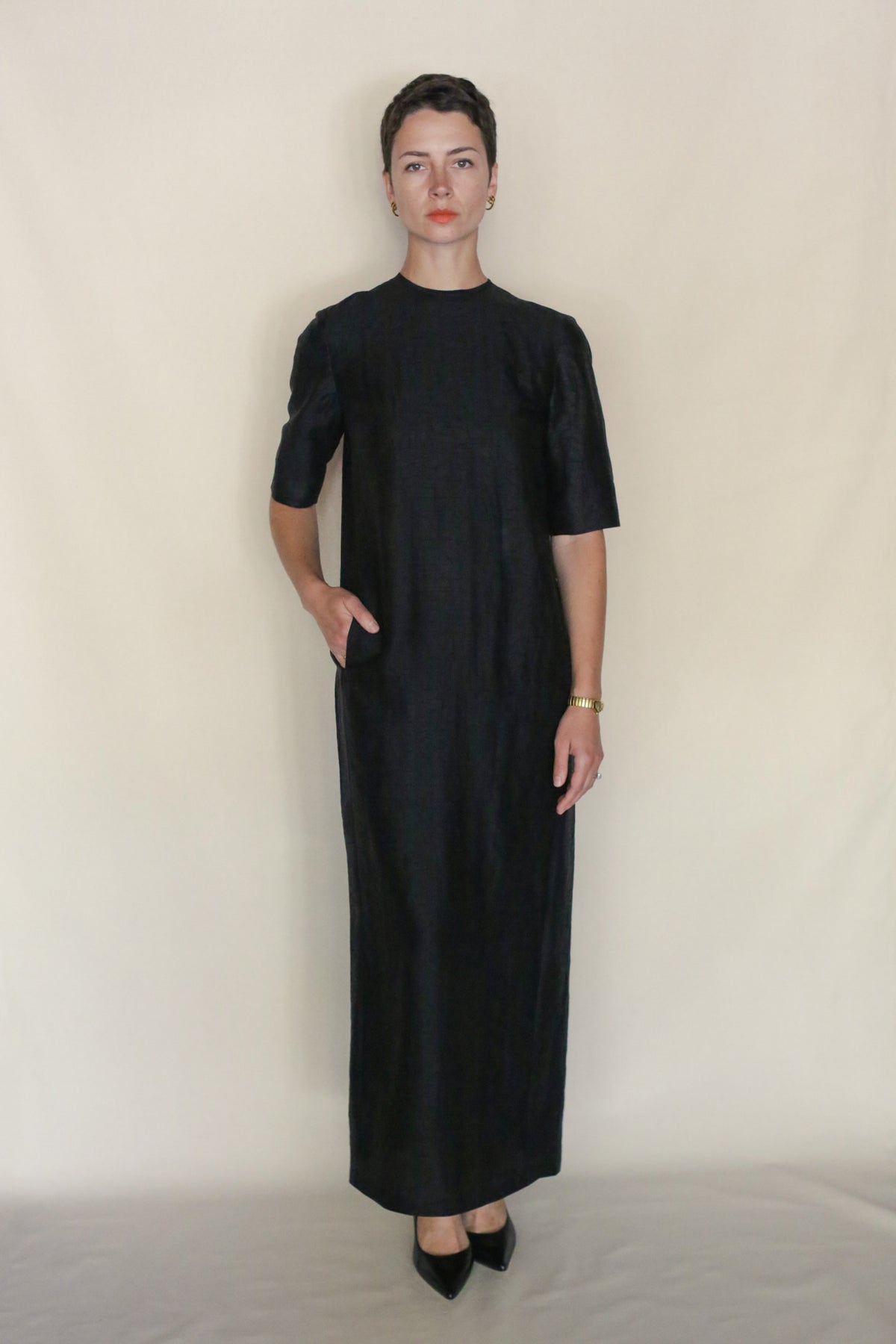 A woman with short dark hair stands against a plain light background, wearing the Earth Mark Dress—a simple, short-sleeved, ankle-length black crewneck dress with pockets. She has one hand in her pocket and wears black heels and a gold bracelet.