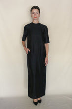 Load image into Gallery viewer, Against a plain light background, a person with short hair and a neutral expression wears the ankle-length Crewneck Dress | Xiang Yun Silk Earth Mark in black, styled with black heels, a gold wristwatch, and one hand in their pocket.