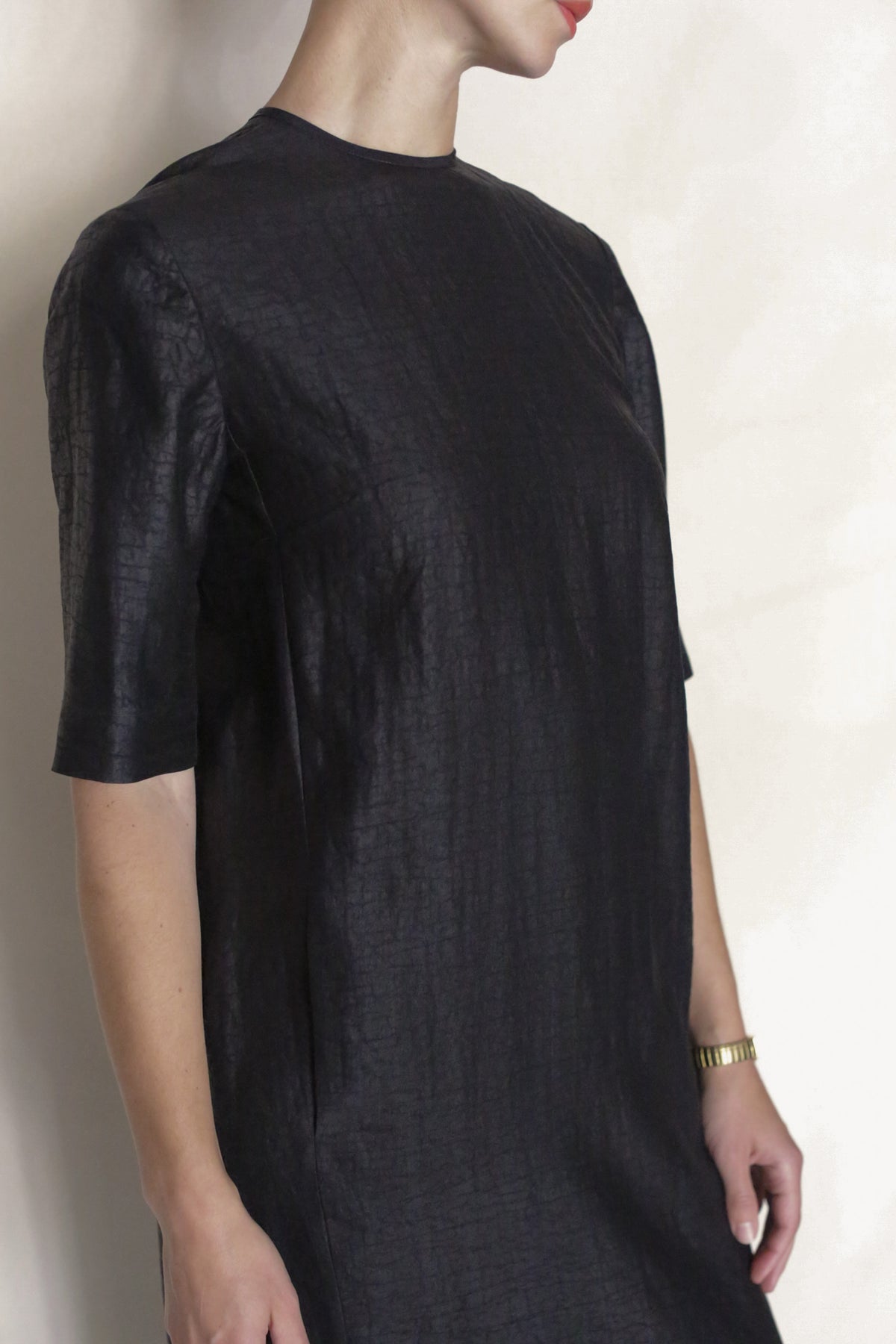 A person wearing the Earth Mark Dress, a black short-sleeved, textured silk dress, and a gold wristwatch stands against a light background. Their face is partially out of frame.