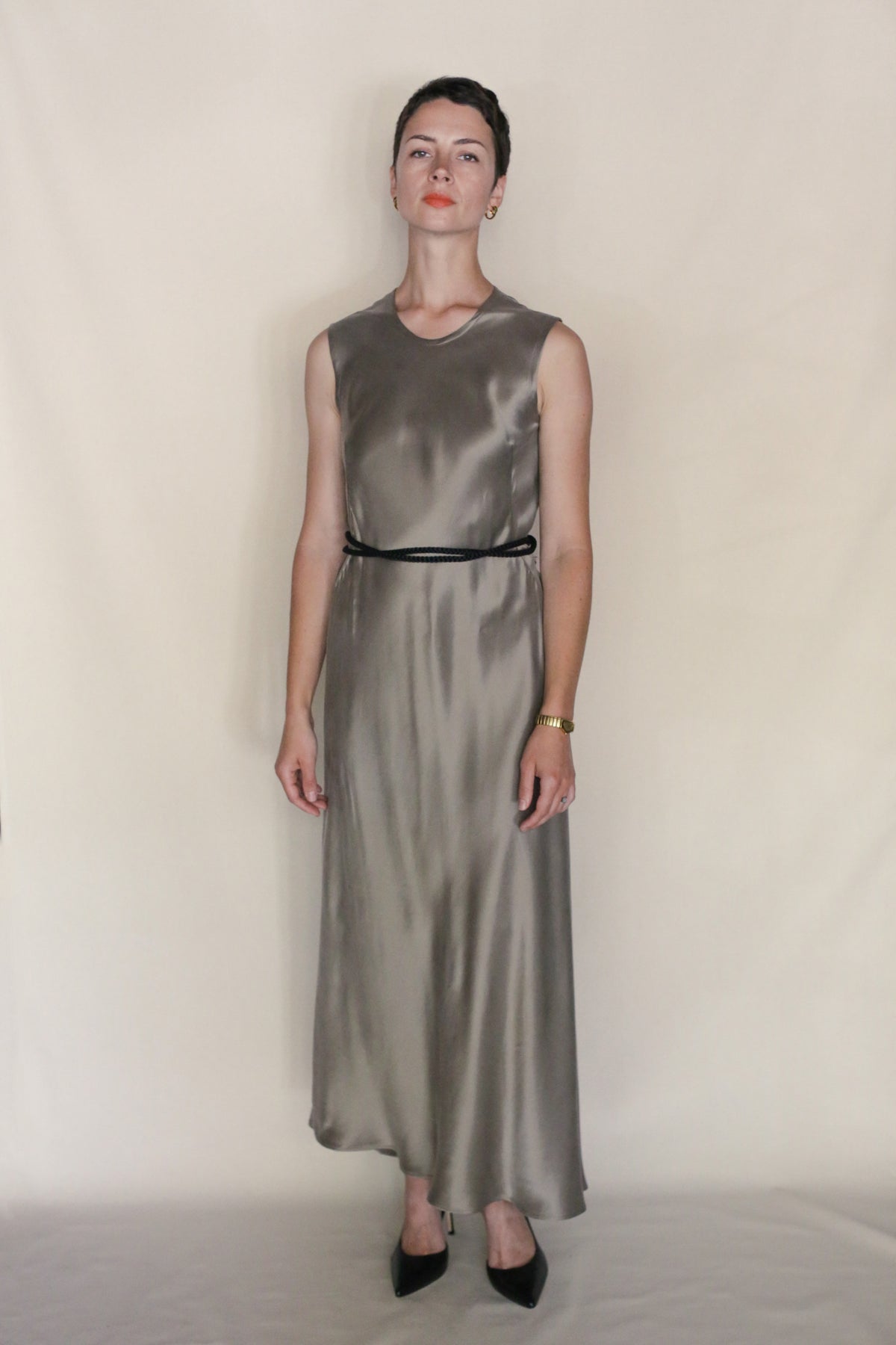 A person stands against a plain light background, wearing the Bias Dress—an ankle-length, sleeveless silver dress styled with a black belt, black pointed-toe shoes, a gold watch, and small earrings.