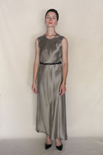 Load image into Gallery viewer, A person with short hair and a calm expression stands before a neutral backdrop, wearing the Bias Dress in Pewter, styled with a black belt, black pointed-toe heels, and a watch.