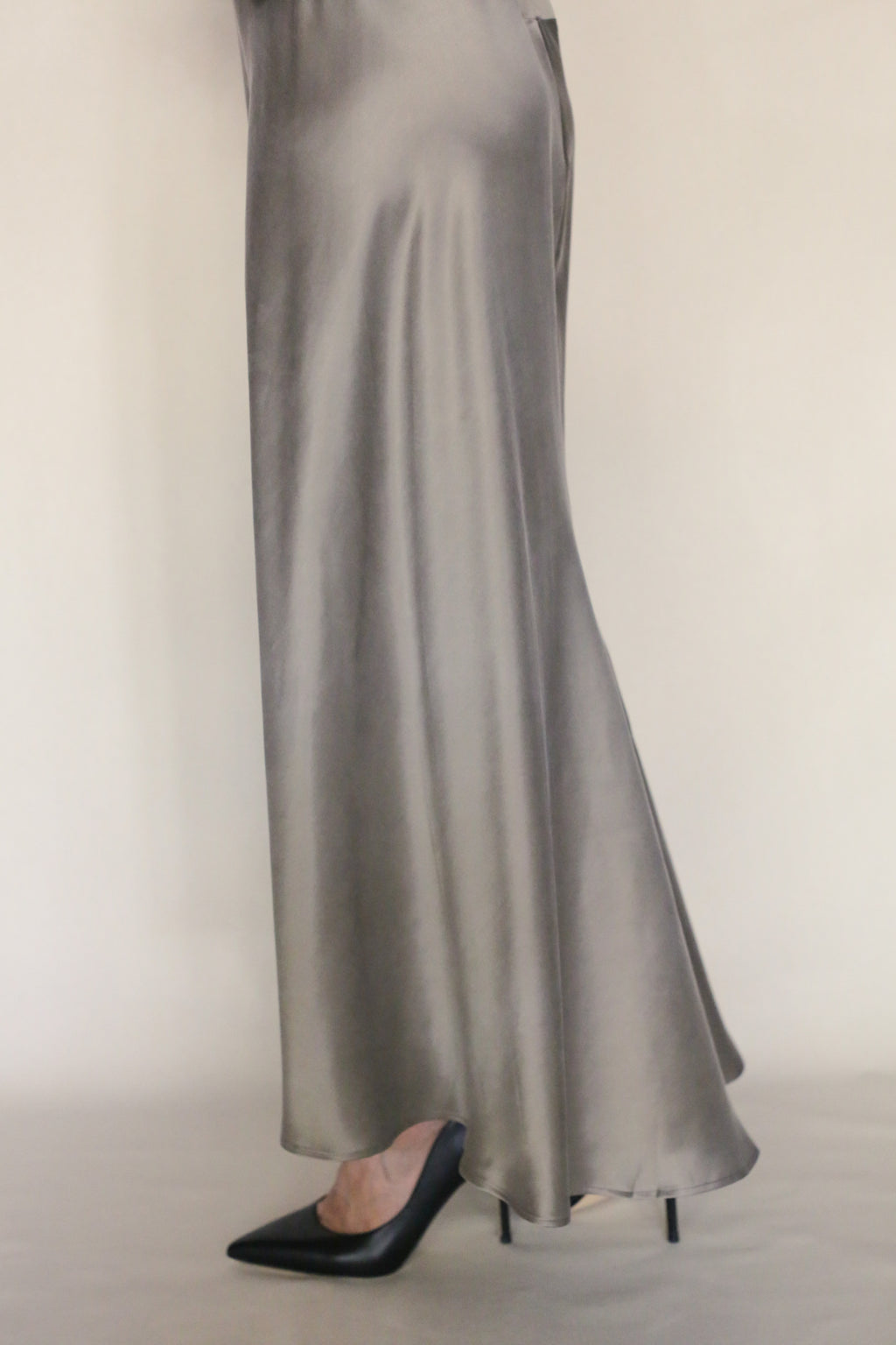 Only the lower body is visible as a person stands against a plain light background, wearing the Bias Dress—a long, flowing silver bias-cut style—with black pointed-toe heels.