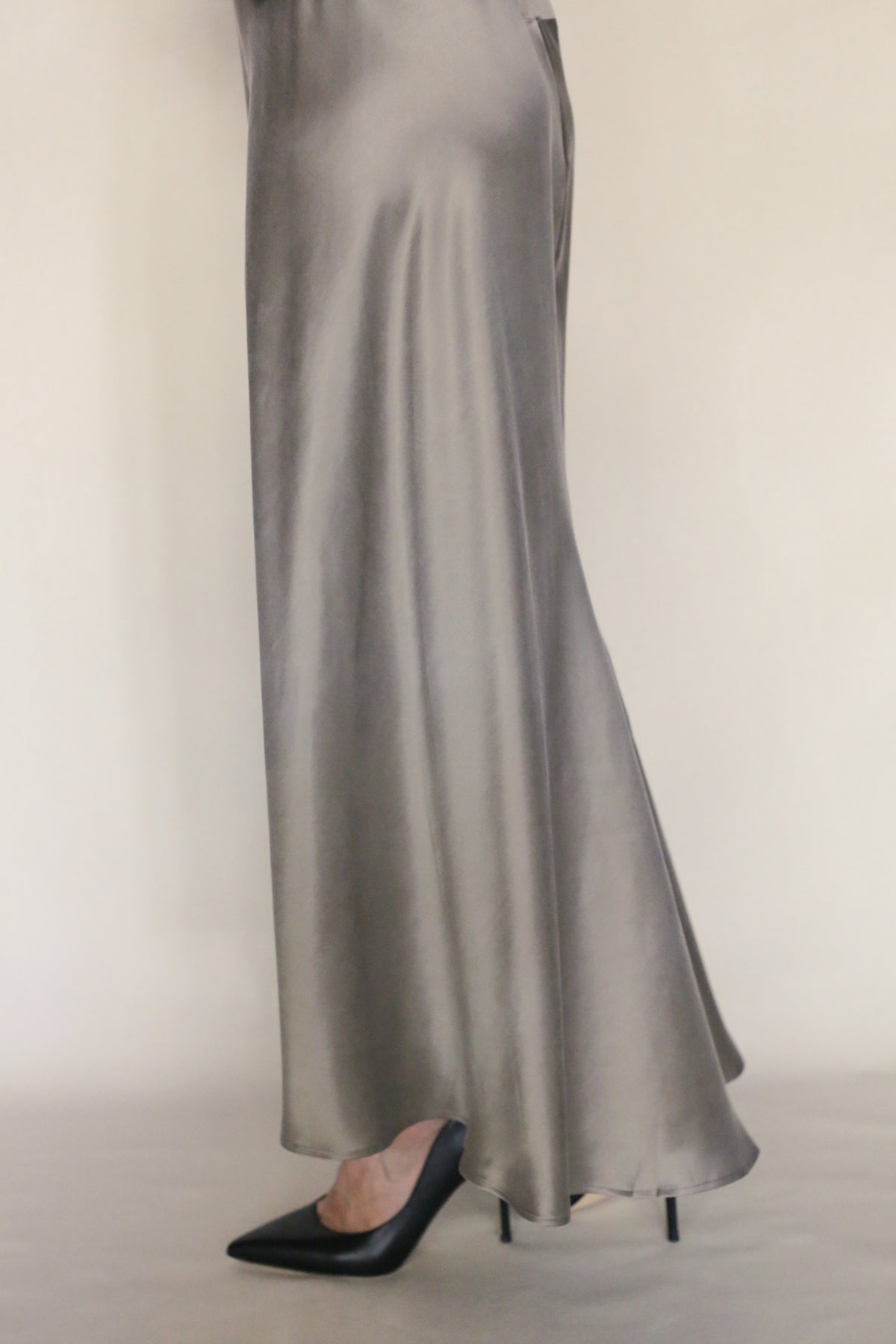 Only the lower body is visible as a person stands against a plain light background, wearing the Bias Dress—a long, flowing silver bias-cut style—with black pointed-toe heels.