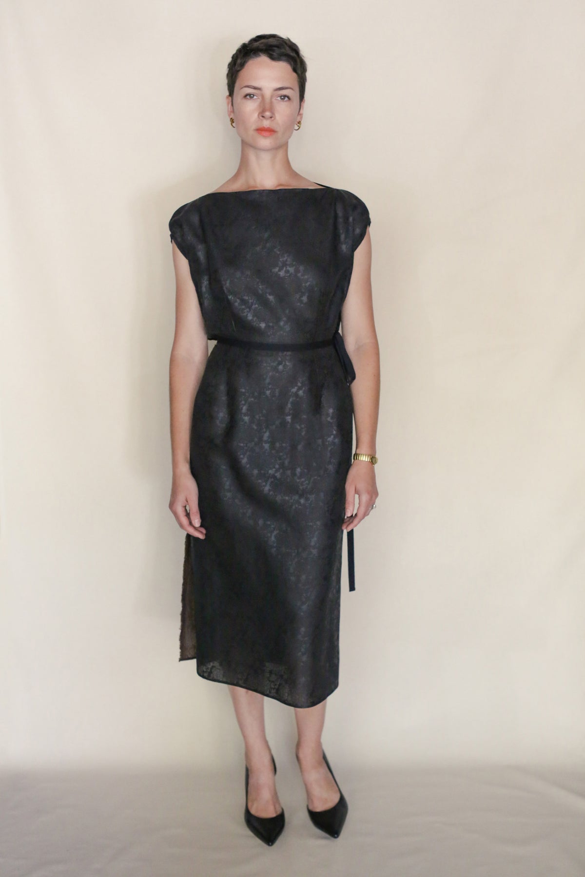 A person with short dark hair stands against a plain light background, wearing a black, knee-length, sleeveless Endless Bloom Futo Dress and black pointed heels, accessorized with a watch and earrings.