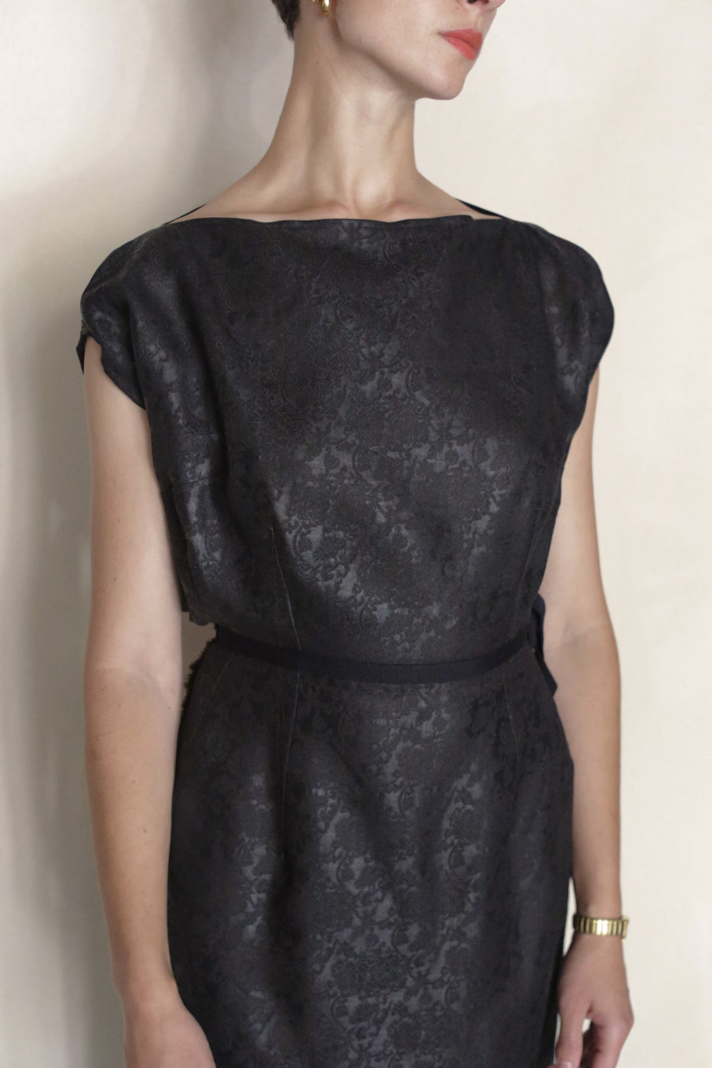 A person wears the black, short-sleeved, patterned Endless Bloom Futo Dress with a gold wristwatch, standing against a plain background. Their face is partially out of frame and turned to the side.