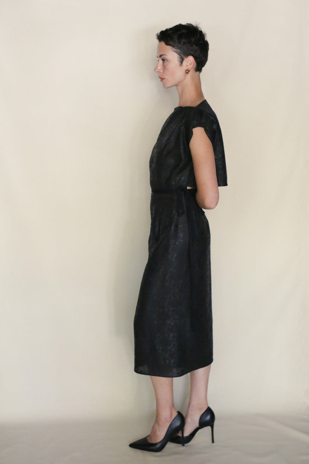 A person with short dark hair stands in profile against a light background, wearing the Endless Bloom Futo Dress—black, reversible, knee-length with short sleeves—and black heels, arms bent behind their back.