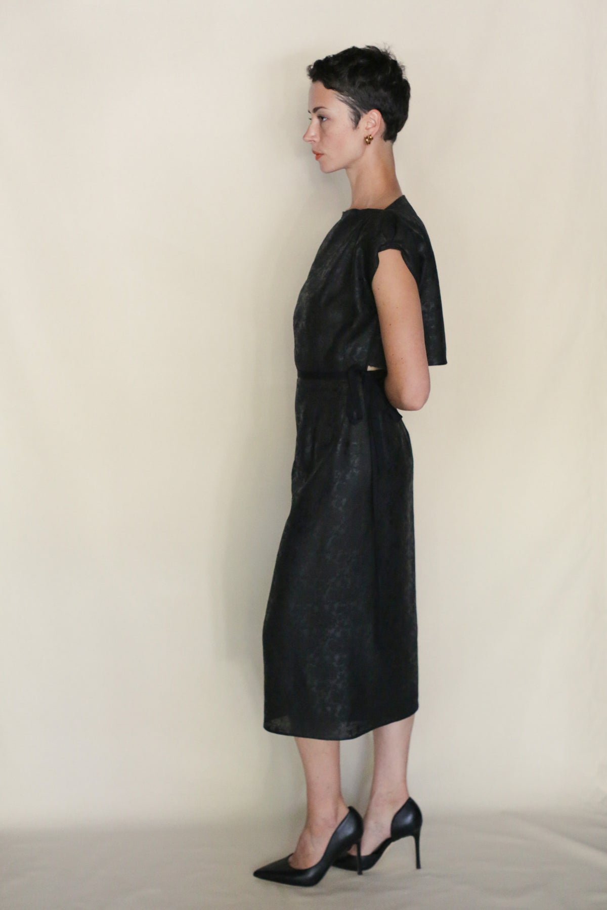 A person with short dark hair stands in profile against a light background, wearing the Endless Bloom Futo Dress—black, reversible, knee-length with short sleeves—and black heels, arms bent behind their back.