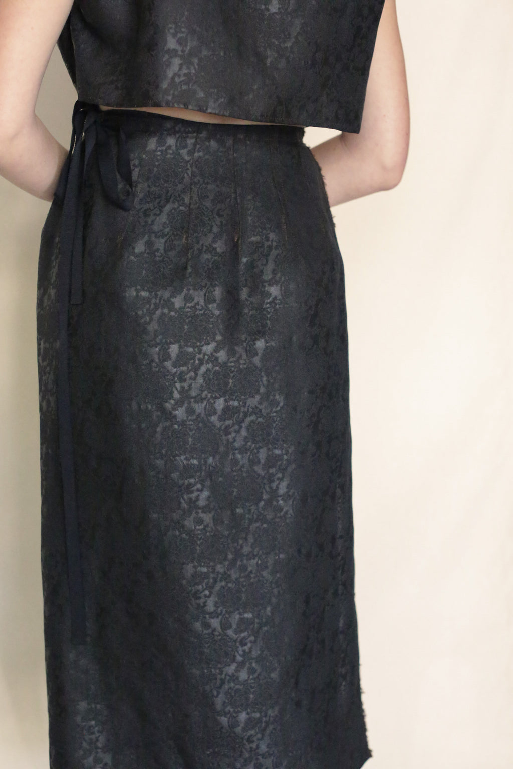 A person wears the Endless Bloom Futo Dress, a dark, textured silk piece styled as a two-piece with a fitted skirt featuring a subtle floral pattern and waist ribbon. The top matches the skirt. Background is plain and light-colored.