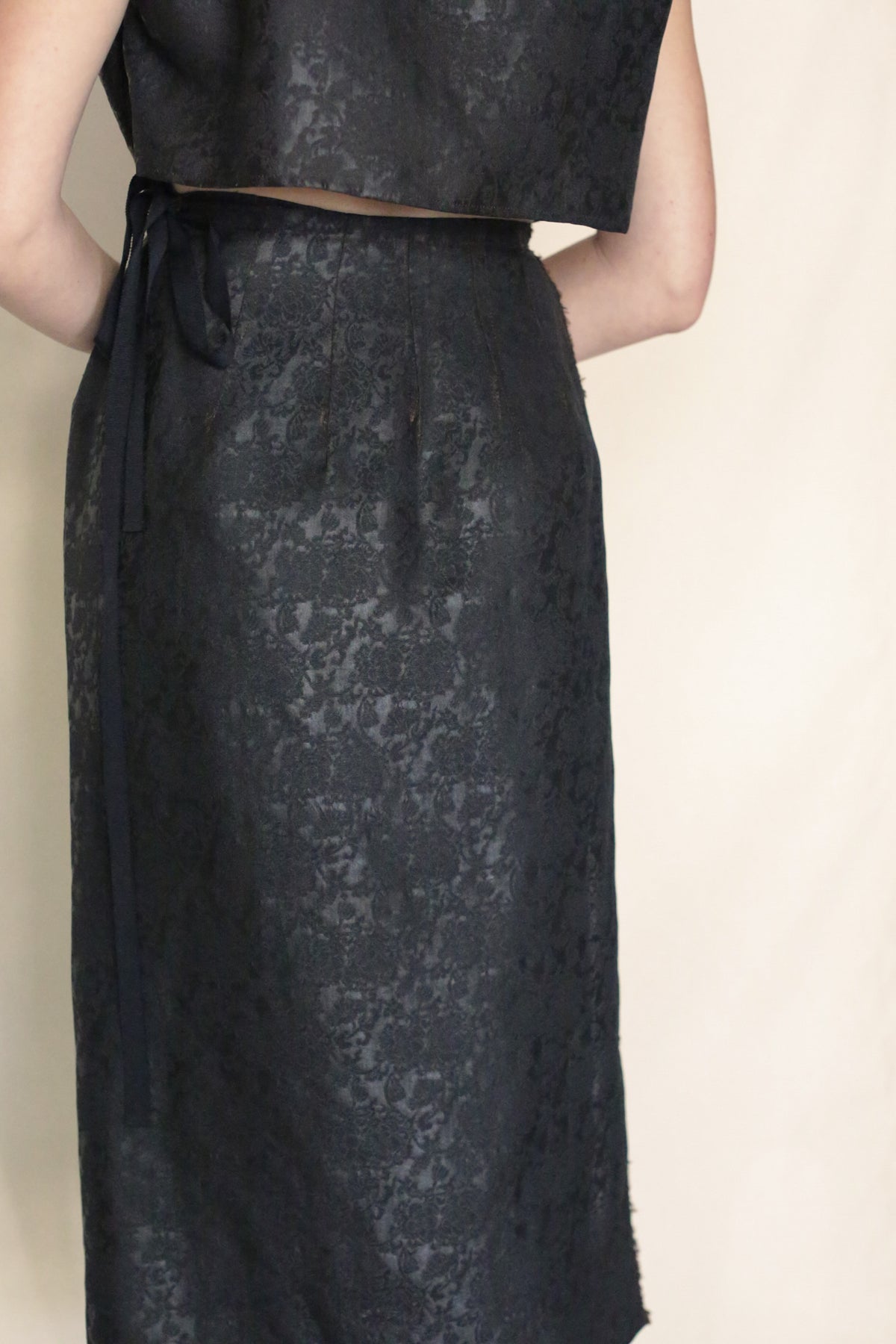 A person wears the Endless Bloom Futo Dress, a dark, textured silk piece styled as a two-piece with a fitted skirt featuring a subtle floral pattern and waist ribbon. The top matches the skirt. Background is plain and light-colored.