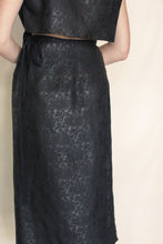 Load image into Gallery viewer, A person wearing the Futo Dress Reversible in Xiang Yun Silk Endless Bloom, showcasing a black textured silk with subtle floral patterns, high waist ribbon detail, and sleeveless top. The background is plain light beige.