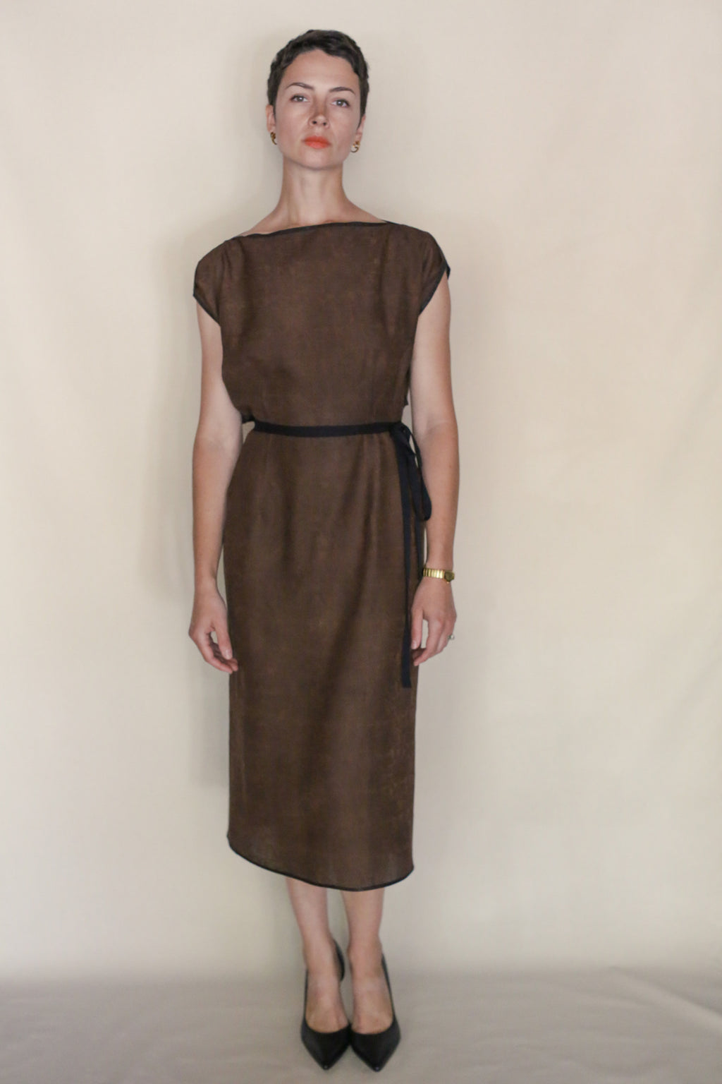 A woman with short dark hair and a neutral expression stands against a beige background wearing the Endless Bloom Futo Dress, accessorized with a black belt, black heels, and a gold wristwatch.