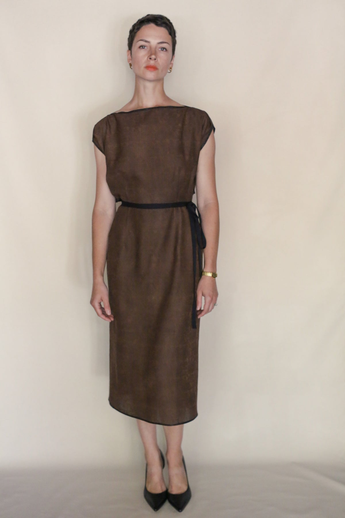A woman with short dark hair and a neutral expression stands against a beige background wearing the Endless Bloom Futo Dress, accessorized with a black belt, black heels, and a gold wristwatch.