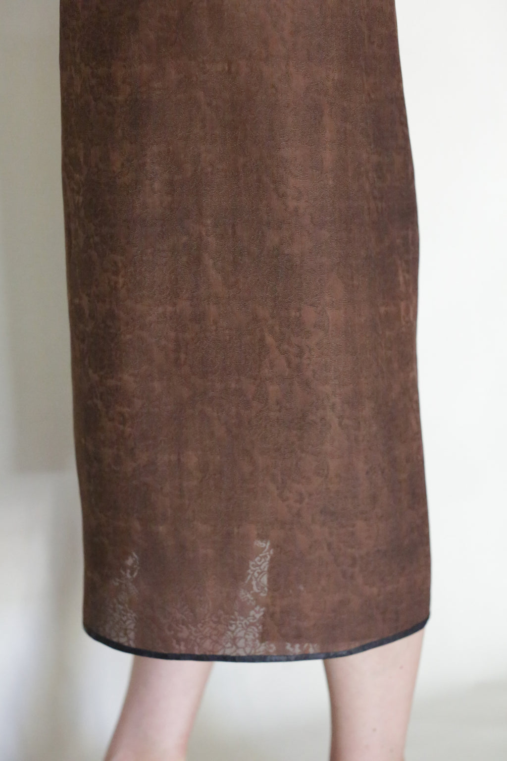 A close-up shows a person wearing the Endless Bloom Futo Dress—a brown, textured, knee-length piece with a subtle pattern—standing against a plain light background. Only the lower part of the dress and legs are visible.